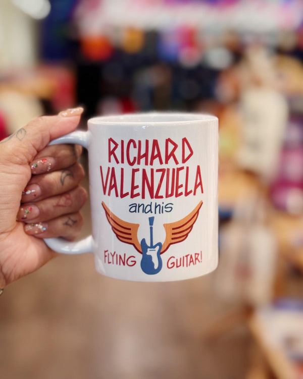 White ceramic mug with vintage-style text that reads 'Richard Valenzuela and his Flying Guitar' with an image of a blue guitar and orange wings from Sew Bonita in Corpus Christi, Texas